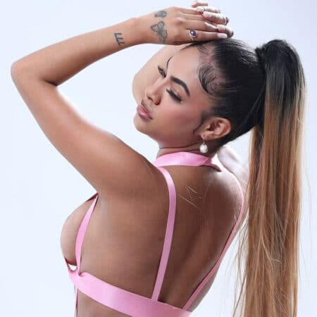 Back view of a woman with a long high ponytail wearing a pink strappy top, arms raised over her head, eyes closed.