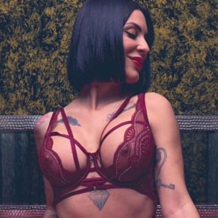 Woman with a black bob wearing burgundy lace lingerie, standing outdoors, smiling to the side.
