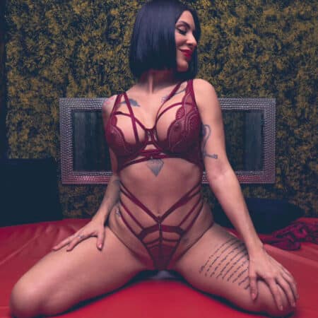 Woman in burgundy lingerie posing on a red bed, visible tattoos on arms and torso, black bob haircut, looking to the side.