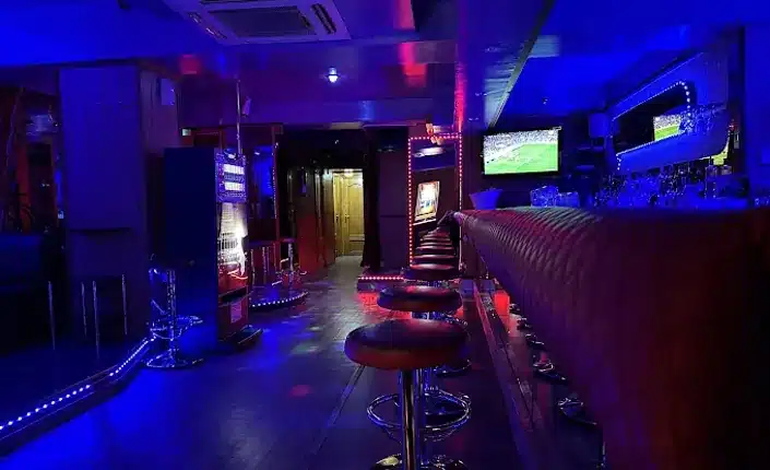 Dimly lit bar lounge with blue neon lighting inside Breston Barcelona, a long counter with red bar stools, and TV screens on the walls showing sports.