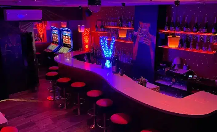 A neon-lit bar inside Pink Lady Barcelona with a curved white counter, red stools, and blue/purple lighting on shelves and decor in the background.