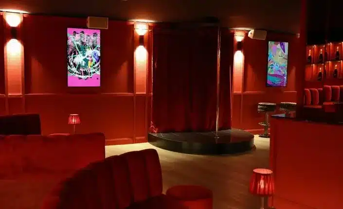 Dim red-lit lounge with a circular black performance pole in the center, red booths, and abstract posters on the walls inside Play Beach Barcelona.